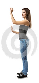 Side view of young woman in top and jeans standing, one arm with open palm at belly level, other before face in pinch