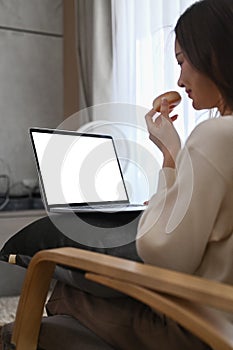 Side view of young woman in warm woolen sweater eating donut and using laptop in cozy living room
