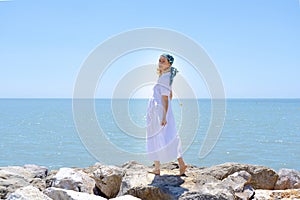 Side view of a young woman wearing a white dress and hair scarf standing on rocks on a sea background against a bright