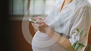 Side-view of youthful woman pregnant women in white t-shirt tapping on smartphone staying in front of the wooden window