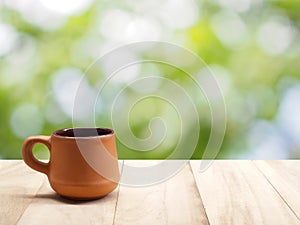 Side of vintage brown baked clay coffee cup on wooden table with natural light bokeh from sunlight between green leaf