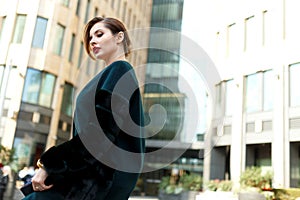 Side virew of a stylish woman walking down the street. Fashionable girl wearing dark green fur coat and clutch.