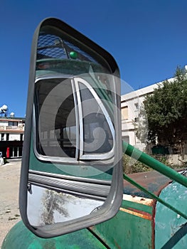 Side windows of an old green rusty vintage bus reflected in the rear view mirror parked in a square in a village in cyprus