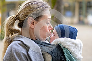 Sideview of a late-term mother in her 40s lovingly holding her newborn baby in a baby carrier and kissing him gently