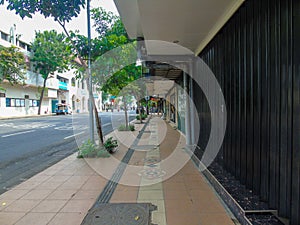 A sidewalk for pedestrians with a few trees, shophouses that are closed and the black door and the main road beside it