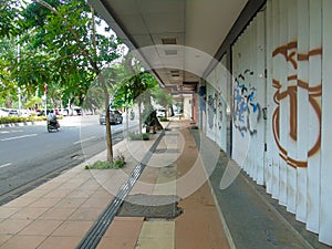 A sidewalk for pedestrians with a few trees, shophouses that are closed and the door gets vandalism and the main road beside it