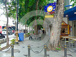 A sidewalk for pedestrians with a few trees, shophouses that are closed and the door gets vandalism and the main road beside it