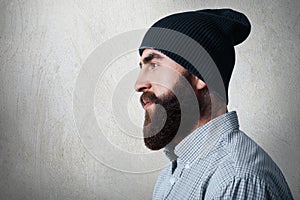 A sideways portrait of stylish bearded man wearing checked shirt and black cap having tatoo on his neck looking into distance whil