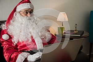 Sideways of Santa Claus relaxing in sofa at home Using laptop for communication and leisure or shopping online.