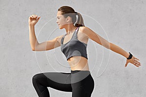 Sideways shot of active woman being in motion, has pony tail, wears sportsclothes, poses against grey background, prepares for run