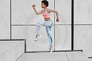 Sideways shot of athletic woman looks ahead, runs up stairs, wants to loose weight, has high jump, wears sportclothes