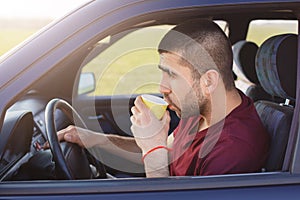 Sideways shot of attractive male driver stops to have break after driving, drinks hot coffee, looks thoughtfully on road while sit