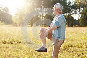 Sideways shot of healthy elderly male stretches legs, has serious expression, wears sport clothes and shoes, stands against nature