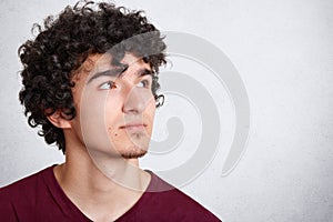 Sideways shot of pensive handsome male with thoughtful expression, looks upwards as notices something, stands against white concre