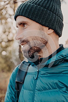 Sideways shot of unshaven man with dark thick beard, wears warm har and anorak, looks pensively aside, poses outside, dreams about