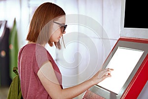 Sideways shot of young Caucasian female with bobbed hairstyle, wears trendy shades, uses ATM machine for withdrawing money,
