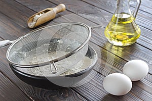 Sieve on black bowl with flour. Glass bottle with butter, eggs, wooden spoon with flour