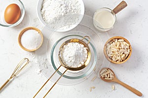 Sifting flour with gold sieve in glass bowl with baking ingredients on marble kitchen table