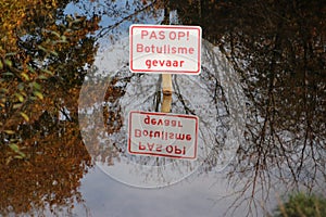 Sign with dutch text Pas op Botulisme gevaar which means Caution, danger for botulism in the water with surface reflection in publ