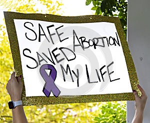 Sign with glitter edge reads Safe Abortion Saved My Live - blurred outdoor background at Reproductive Justice Rally