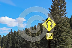Sign beside mountain road in front of evergreen trees warning of extreme curve and speed limit of 20 mph - selective focus
