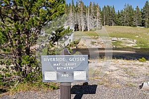 Sign noting times for when Riverside Geyser in Yellowstone National Park might erupt. Geyser is erupting in background