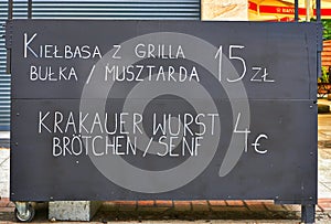 A sign outside a restaurant that reads Krakow Sausage with Buns and Mustard in Polish and German text