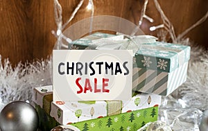 A sign that reads CHRISTMAS SALE stands on the table with gift boxes in the background. Christmas background with decorations from