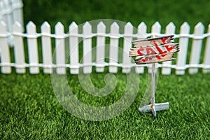 A sign for sale on a green lawn in front of a white fence