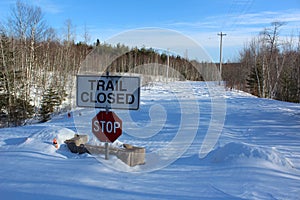 A sign saying trail closed above a stop sign on a snow covered road at a wash-out bridge in the forest