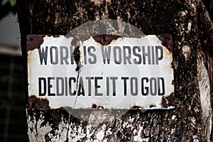 Sign with the words `work is worship dedicate it to god` ruined and hanging from a tree in Rishikesh