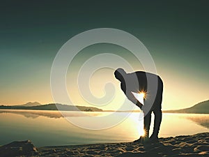 Silhouette of active man exercising and stretching on the lake beach at sunrise.