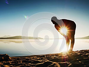 Silhouette of active man exercising and stretching on the lake beach at sunrise.