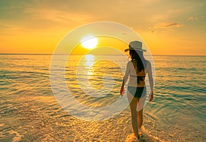 Silhouette adult woman walking at tropical sea with beautiful sunset sky at paradise beach. Happy girl wear bikini and straw hat