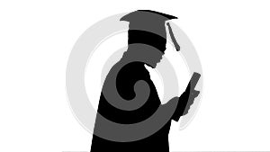 Silhouette African american male student in graduation robe texting on the phone while walking with his diploma.
