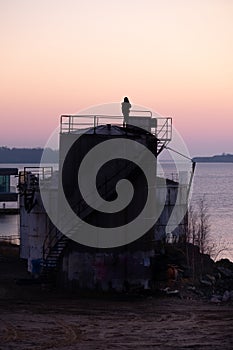 A silhouette of an alone man standing on top of an abandoned oil storage tank during the sunset