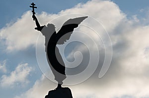 Silhouette of an angel with a cross in his hand on a background of dramatic sky and sun. Fragment
