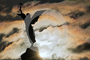 Silhouette of an angel with a cross in his hand on a background of dramatic sky and sun. Fragment of the Monument Mermaid ,