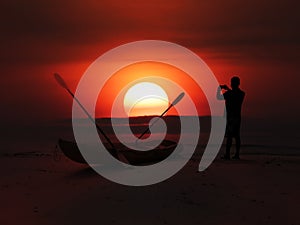 Silhouette of asian man stand and take sunset photo on beach near kayak in evening