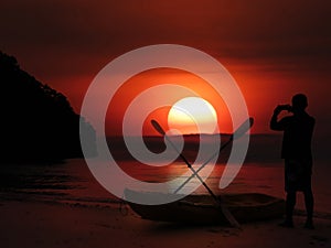 Silhouette of asian man stand and take sunset photo on beach near kayak in evening