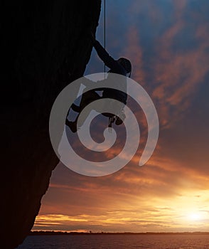 Silhouette of athletic person in safety harness. Climber on rope rock climbing on challenging cliff over water at sunset