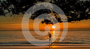 Silhouette back view of woman watching beautiful sunset at tropical paradise beach. Happy girl wear bikini and straw hat