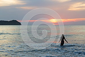 Silhouette back view of young woman in bikini entering into the Pacific Ocean during a pink sunset