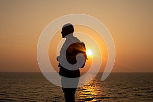 Silhouette of bald vacationer in sunglasses with backpack standing sideways against seascape on seaside at sunset.