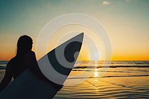 Silhouette of beautiful sexy surfer female with surfboard on the sandy beach at sunset.