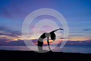 Silhouette of a beautiful woman practicing yoga asana on the beach with sunrise on twilight blue vibrant sky