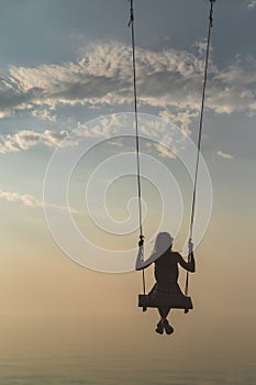 Silhouette of a beautiful young girl who is swinging on a swing against the background of the sky and sea waves