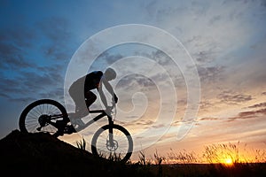 Silhouette of bicyclist wearing helmet and sportswear on mountain bike coming down the hill at sunset
