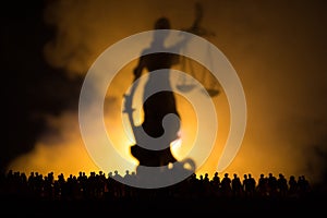 Silhouette of blurred giant lady justice statue with sword and scale standing behind crowd at night with foggy fire background. at