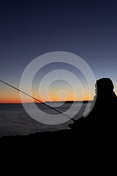 Silhouette of boy with fishing rod at calm sunset fishing off the cliff long exposure photo